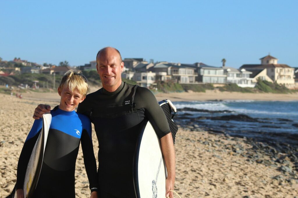 Father and son in wetsuits with surfboards on beach.